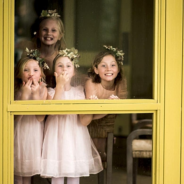 four young girls in pink dresses and flower crowns smile out through a rain-drenched yellow framed window