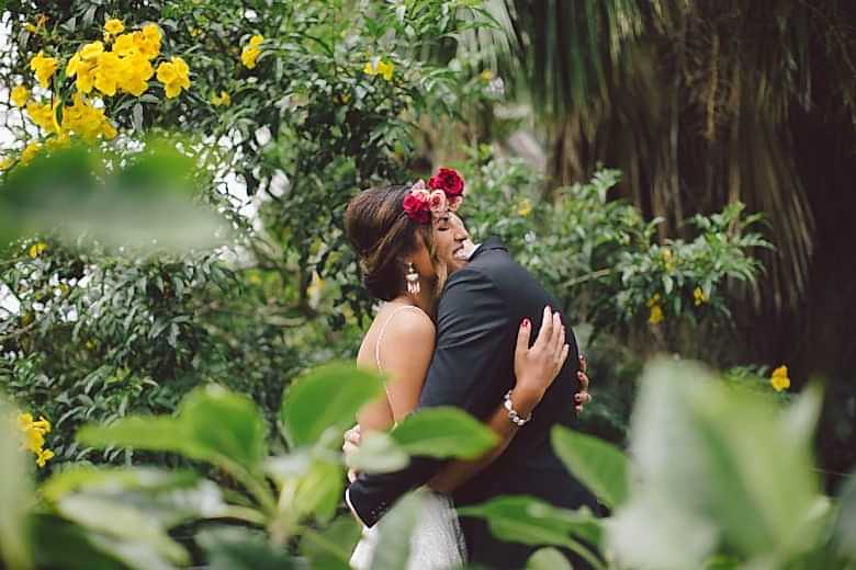 Bride with red and pink flower crown smiles and embraces groom in a lush tropical environ