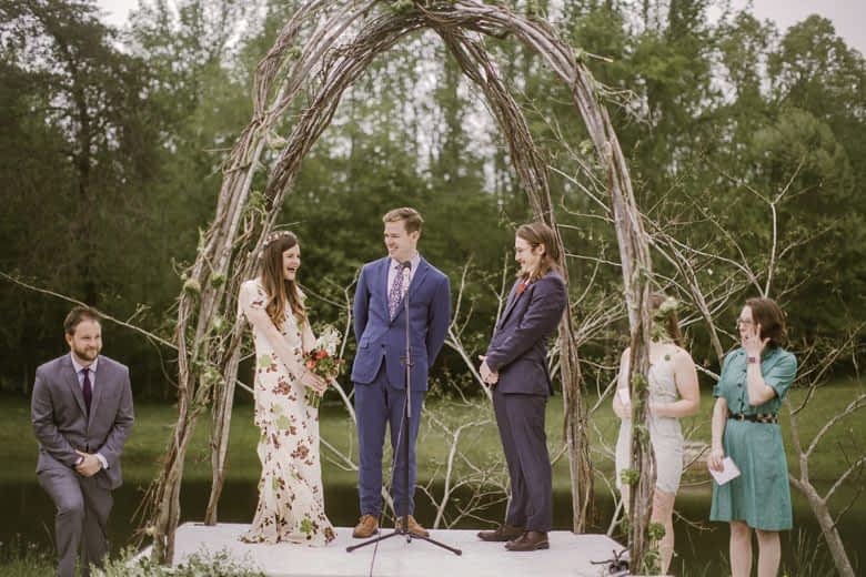 couple in wedding attire (one in a floral dress) stand with officiant under twig and branch arches