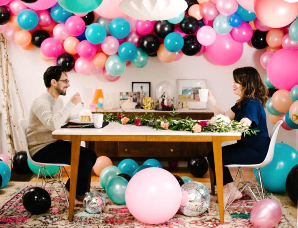 couple sitting at a breakfast table eating wedding cake in a room filled with disco balls and brightly colored balloon installation