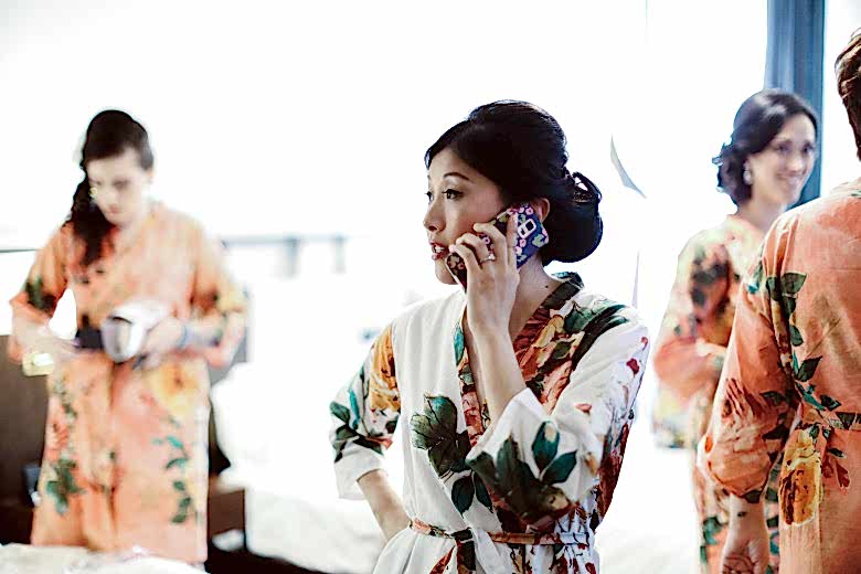 an asian bride wearing a floral robe and a modern updo talks on her cell phone while getting ready for her wedding