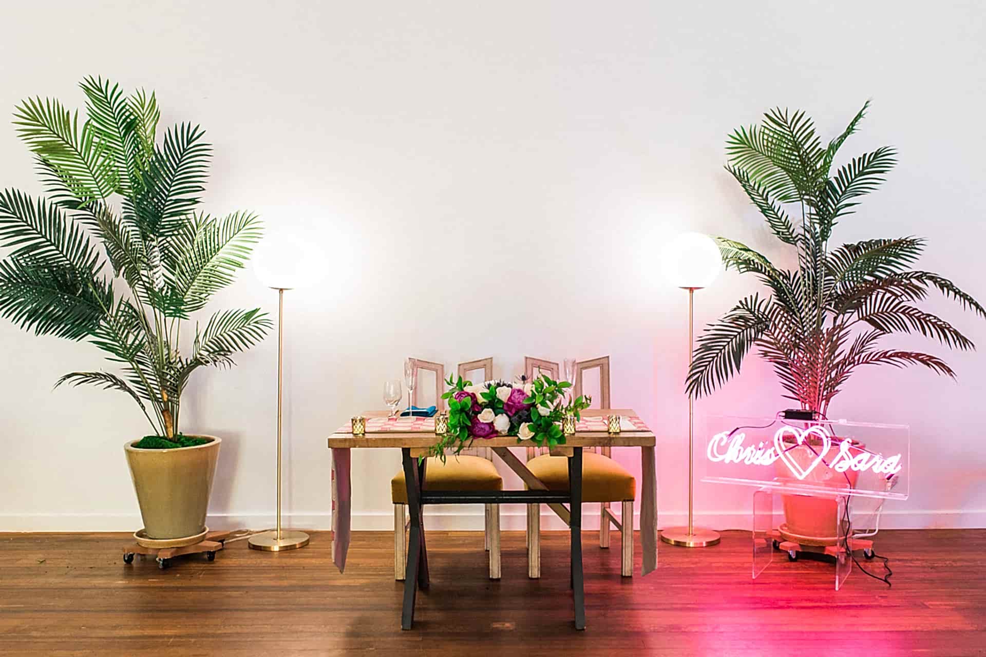a sweetheart table sits in a white room with wood floors. There are two potted tropical plants on either side of the table and a neon sign that says "Chris <3 Sara"