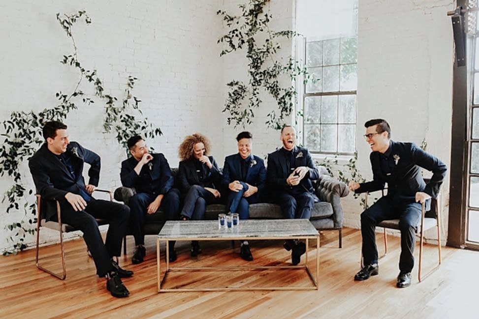 a mixed gender wedding party sits in a minimal room with modern furniture wearing black suits