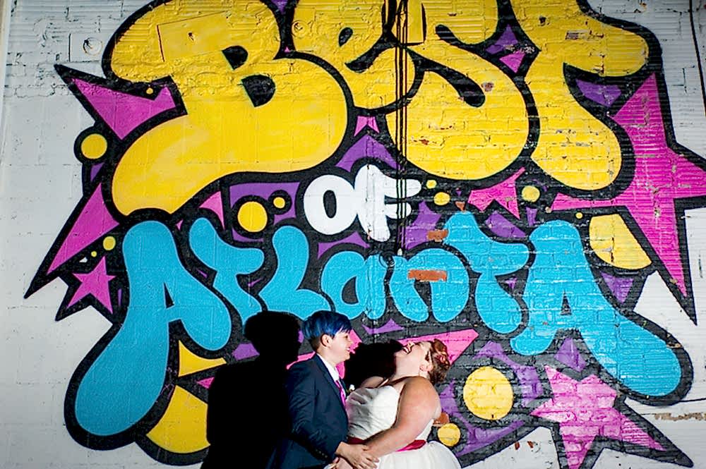 Queer wedding couple holding hands and laughing while standing in front of colorful "Best of Atlanta" mural