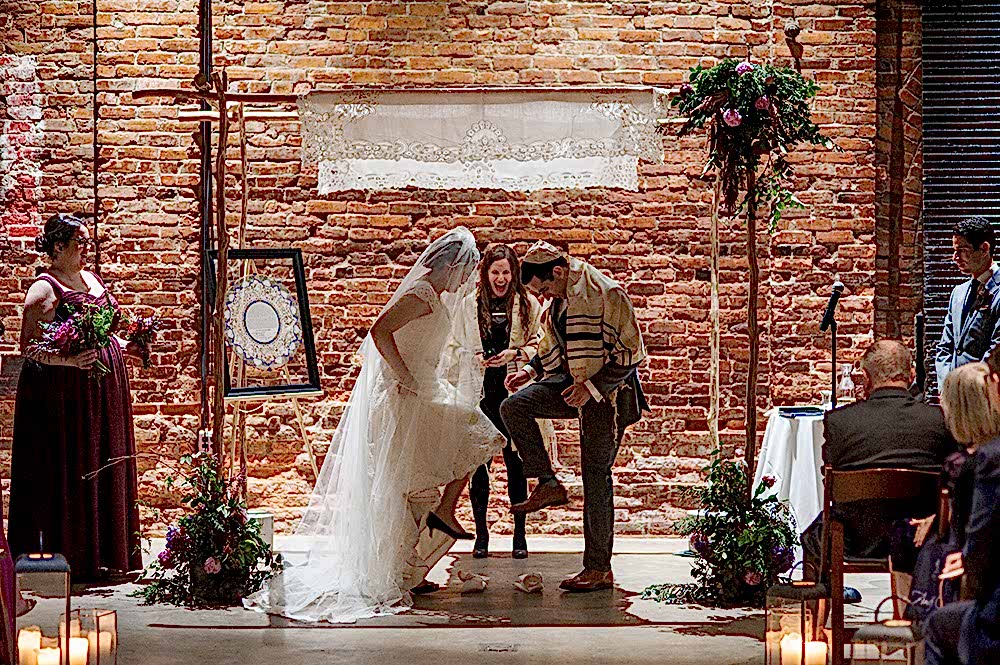 Bride and groom stomp on glasses at altar in front of an exposed brick wall in a photo by You Are Raven