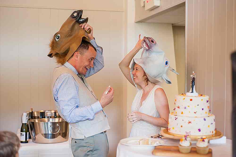 Bride and groom take off silly horse heads and grin at each other during their cake cutting in a photo by You Are Raven