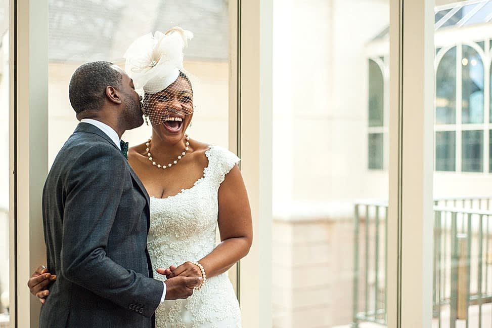 A black bride in a birdcage veil and lace gown laughs joyously as her groom whispers in her ear in a photo by You Are Raven