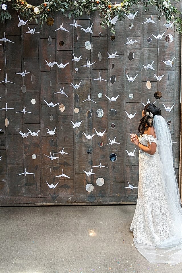 a bride stands in front of paper cranes and planet cutouts hanging vertically in front of a cement wall, bordered on top with a greenery garland
