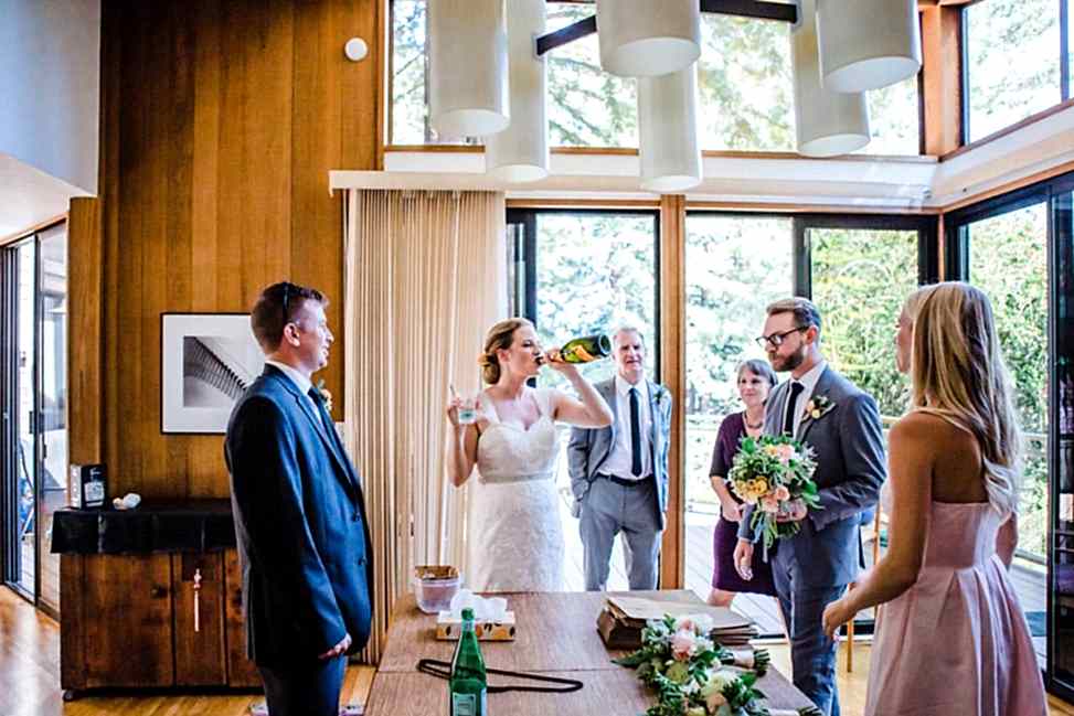 people standing around a table watching woman in a wedding dress drink champagne from the bottle