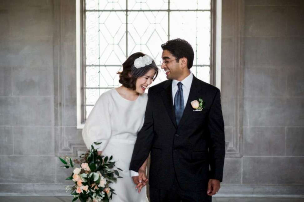 a woman in a wedding dress laughs at a joke by a man in a suit inside San Francisco City Hall