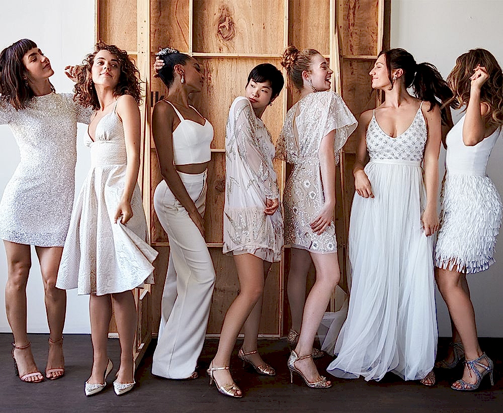 A row of women posing in wedding dresses with five women in short wedding dresses, one in a long dress, and one in a pants separate with a crop top