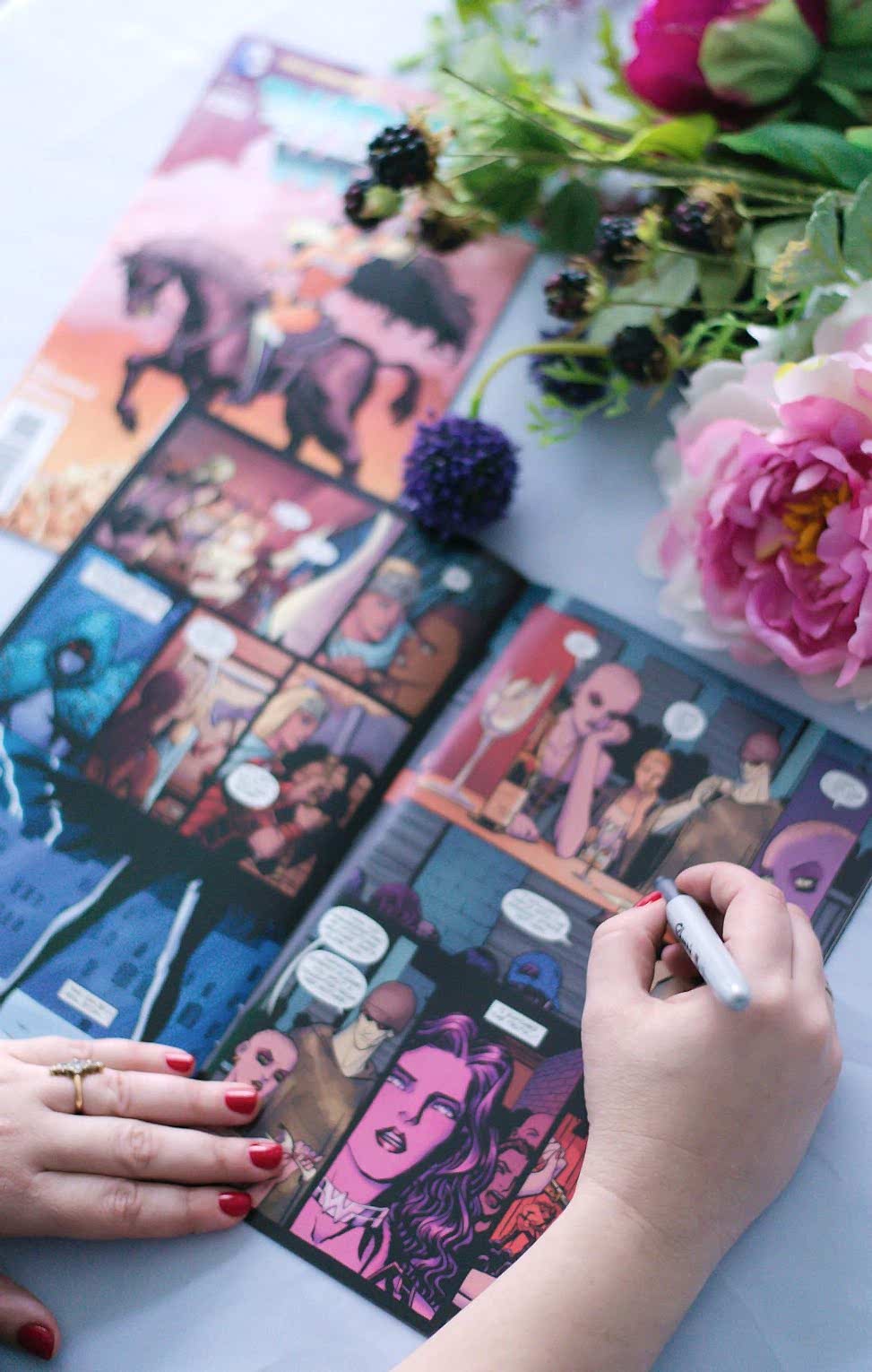 a woman signing a comic book on a table surrounded by flowers