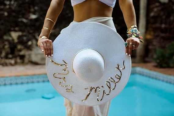 A woman in a white pool wrap with lots of bracelets stands in front of a pool holding a white sunhat with "Mrs. Miller" written in gold sequins on the brim.