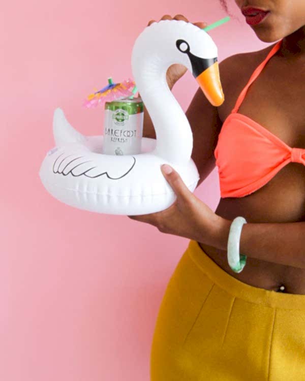 Woman in a brightly colored bikini top drinks Sparkling Wine from a can with a cocktail umbrella with a straw. The can is on an inflated swan pool drink holder.