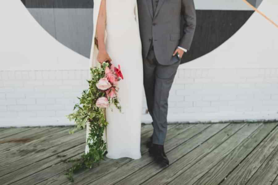 a person in long white dress holds a cascading bouquet alongside as she stands next to a person in a grey suit