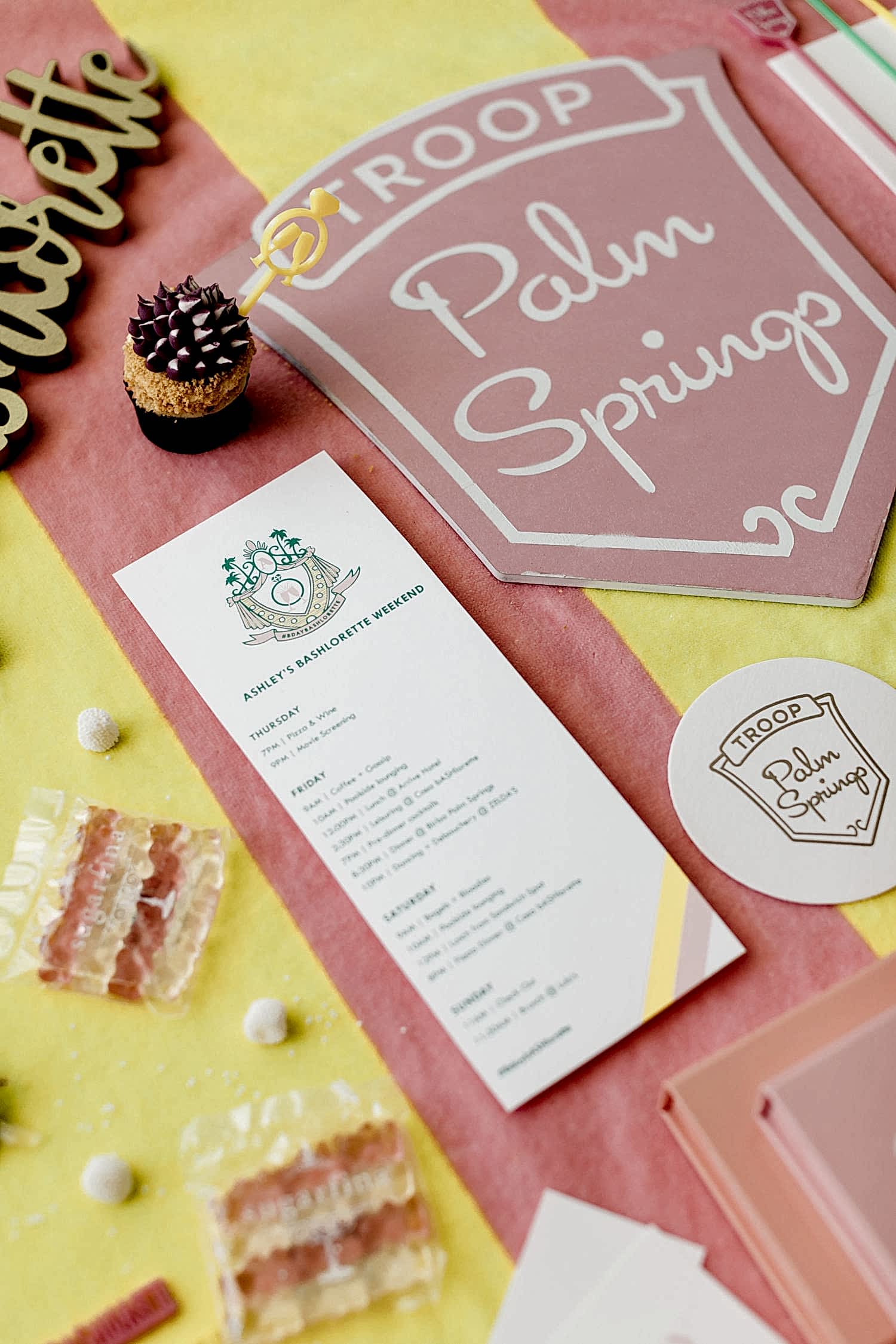 Troop Palm Springs pink, yellow, and white decor and printed collateral, including itinerary, mini cupcake, candies, and other bachelorette accoutrement