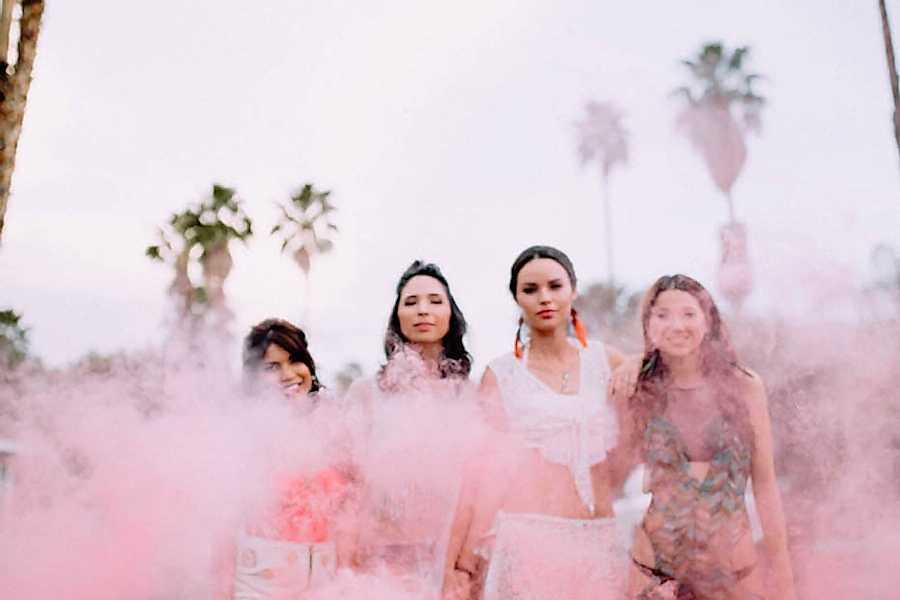 a group of women in sundresses stand in front of palm trees, engulfed in atmospheric pink smoke