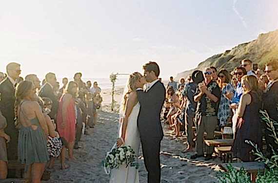 Two people kiss in front of a hundred other people, at the beach.
