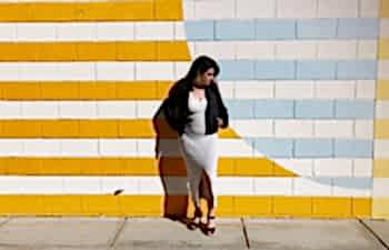 A woman leans against a yellow, white, and light blue striped wall wearing a leather jacket and white ankle-length sundress