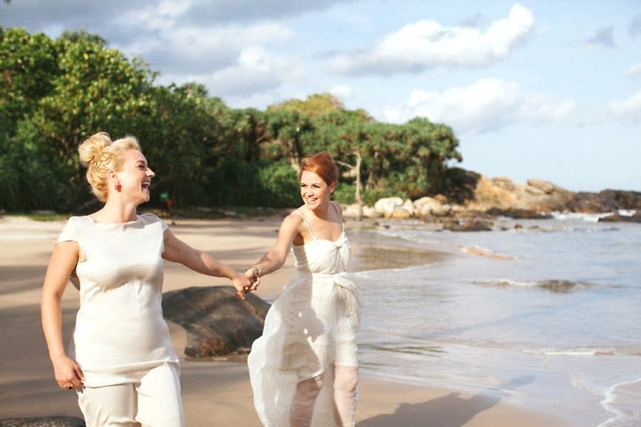 Two women in wedding dresses hold hands on a beach, smiling