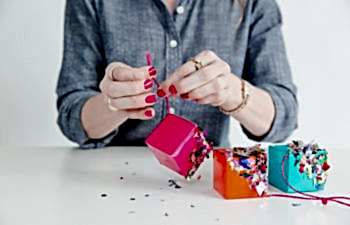 brightly manicured woman in a chambray shirt crafts with small bright cubes half covered in confetti