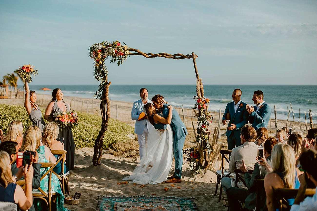 Two people kiss while other people cheer, at the beach.