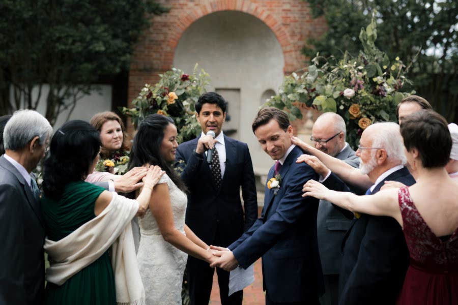 Couple holding hands at wedding ceremony in front of officiant, surrounded by family placing hands on them