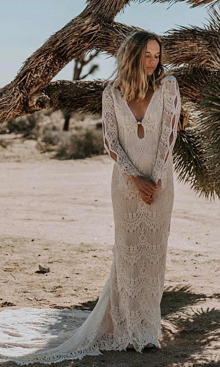 A woman wears an off-white crochet-style lace dress with long sleeves, a plunging neckline that fastens at the bust, and a train