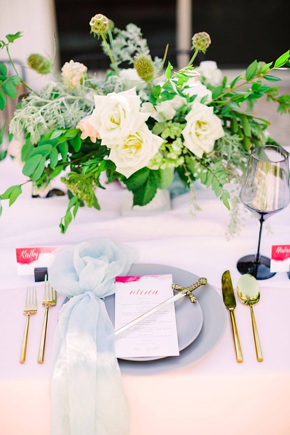 game of thrones inspired placesetting with pink tablecloth and blue silk napkins with palm tree candleholders and sword letter openers