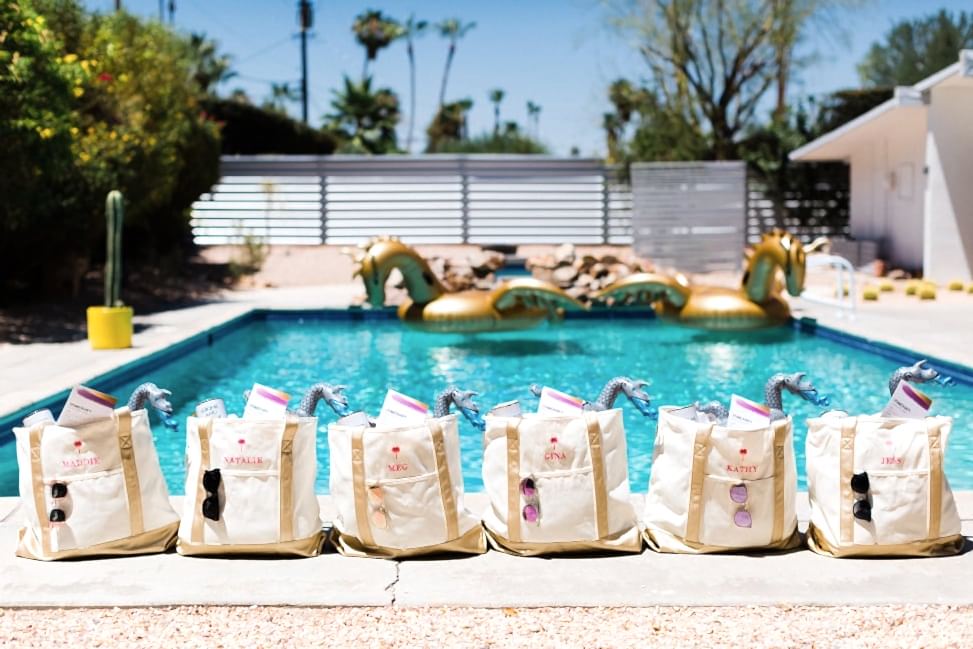 personalized embroidered tote bags with sunglasses, weekend itineraries, and inflatable dragon pool cupholders in front of a pool with gold dragon floaties