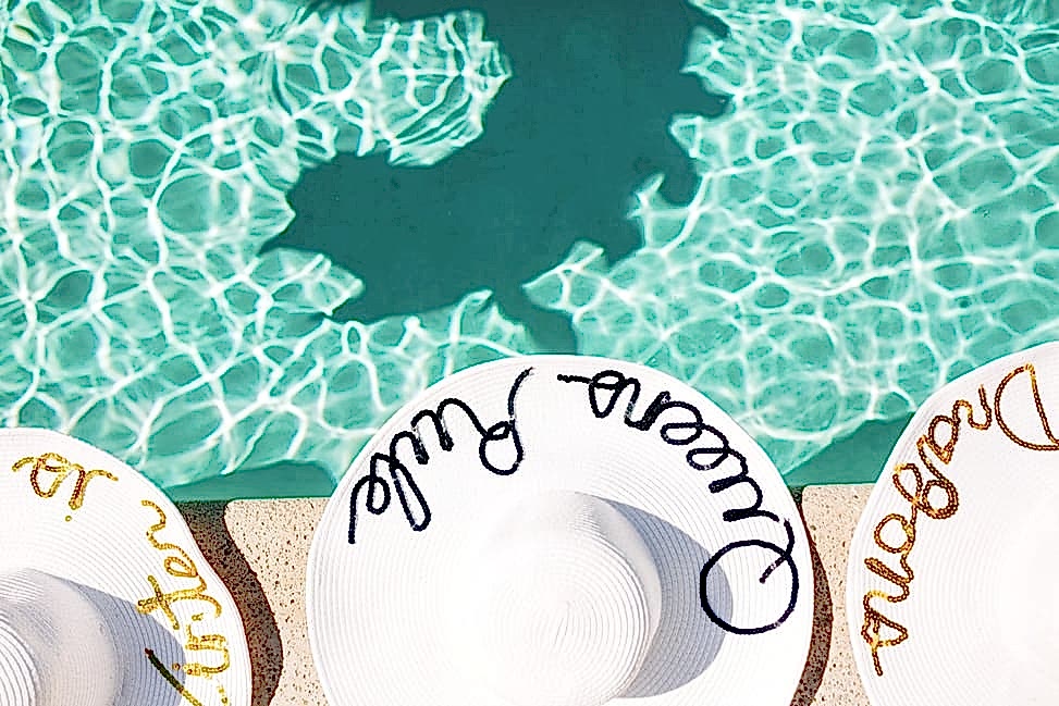 Three sun hats at the edge of a pool—the middle one reads "Queens Rule" and there is a shadow of a dragon on the bottom of the pool