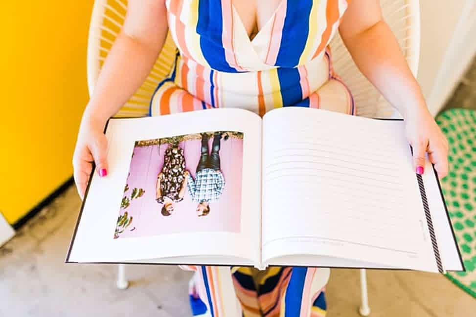 a woman in a striped jumpsuit holds a custom personalized guest book open to a page with an image of a couple in front of a pink wall, opposite a blank lined page