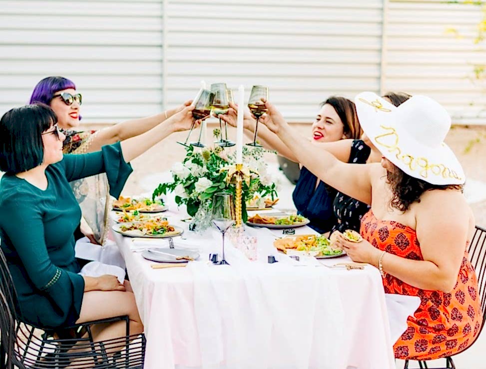Women toasting at a table with a game of thrones inspired tablescape with pink tablecloth and blue silk napkins with palm tree candleholders and sword letter openers