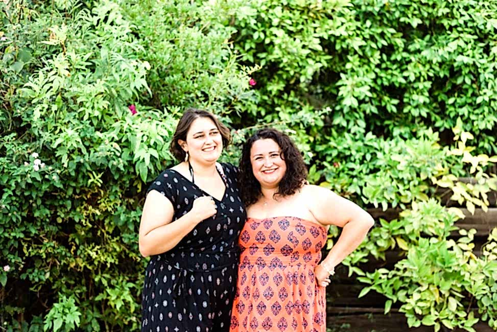 two women stand near each other in front of green bushes, smiling in sundresses