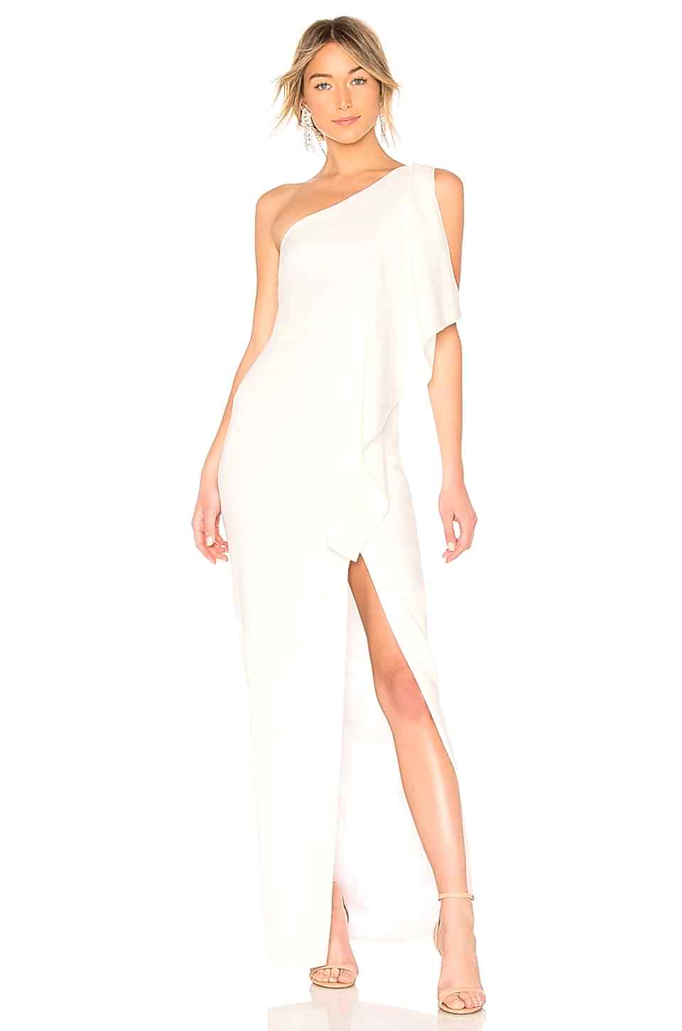 A woman wears a one-shoulder white dress with a large vertical ruffle descending from the shoulder and a high front side slit