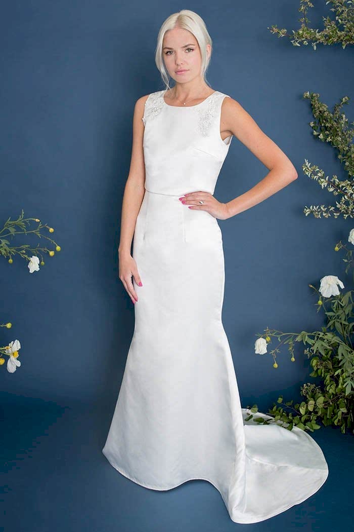 A woman wears a high scoop-neck sleeveless white dress with white appliqué on the shoulders and mermaid skirt with small train