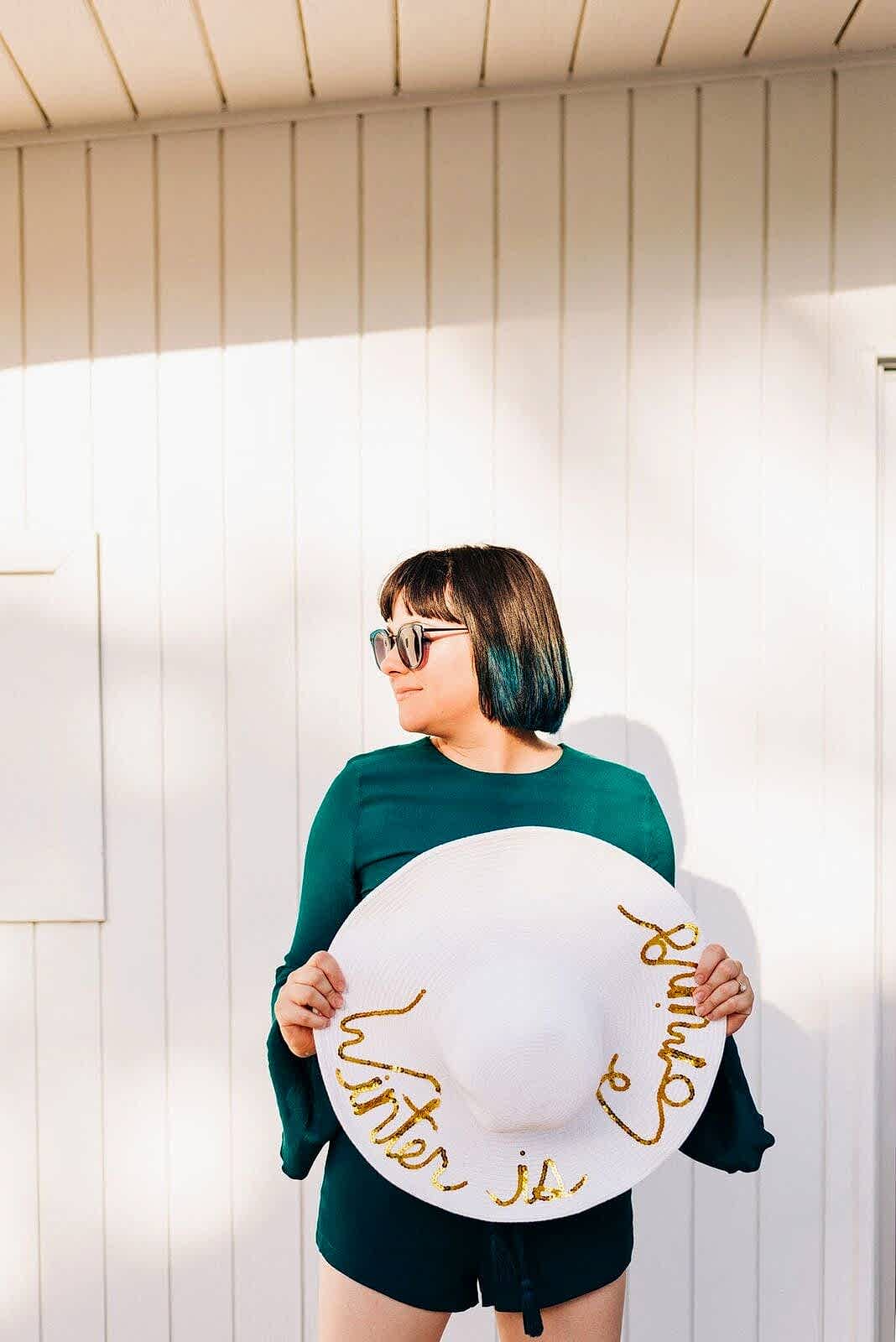 a woman in a green romper wears sunglasses, looks to the side, and holds a white sunhat that reads "Winter is Coming" in Gold