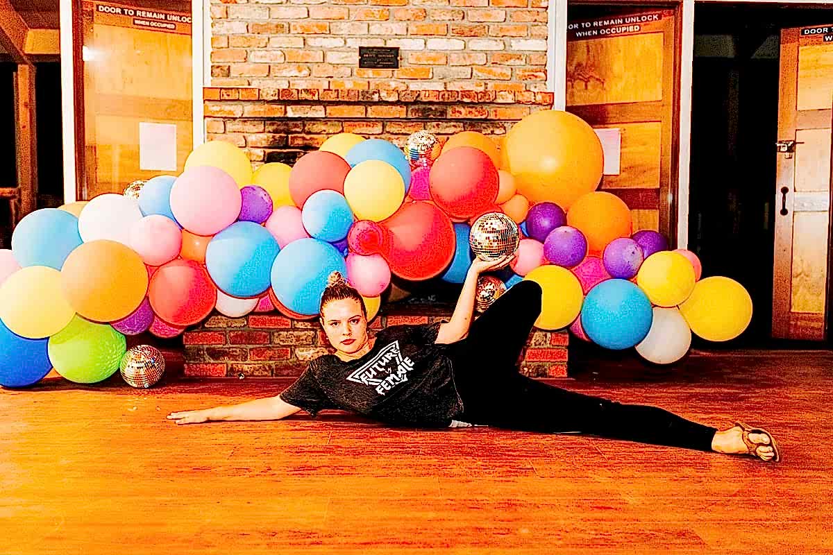 Jess Rutherford, lying in front of a fireplace covered with a balloon installation she made, holding a disco ball, wearing a Future is Female teeshirt at the compact summer camp in la Honda, CA