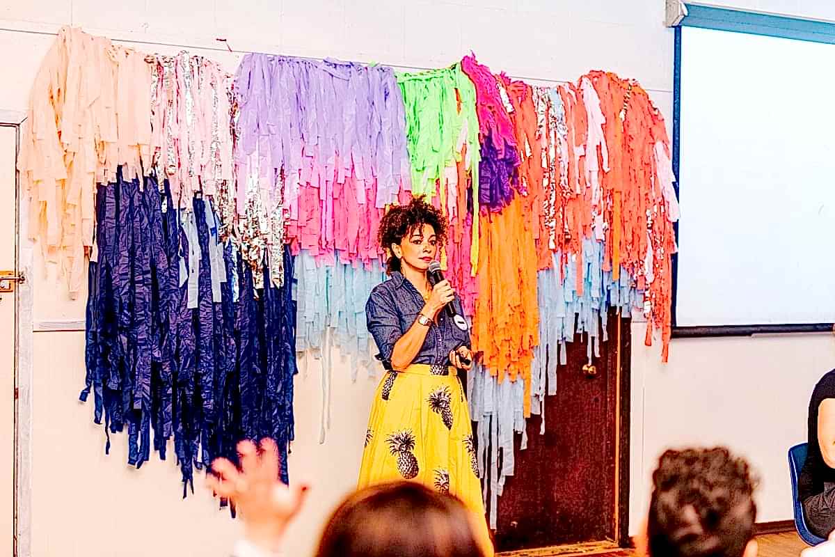 Cyndie Spiegel, wearing a pineapple skirt, standing in front of a colorful fringe installation, holding a microphone during her keynote at the compact summer camp in la Honda, CA