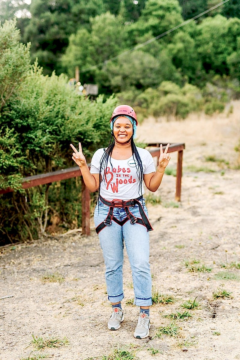 A woman with long braids wearing a babes in the woods tee shirt and zip lining harness and helmet, smiling and throwing peace signs amid the redwoods at the compact summer camp in la Honda, CA
