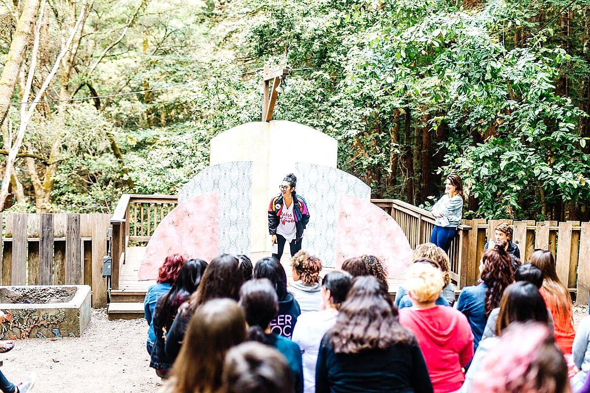 A woman in front of the sunrise installation on the wood deck laughing and talking to a large group of women among the redwoods at the compact summer camp in la Honda, CA
