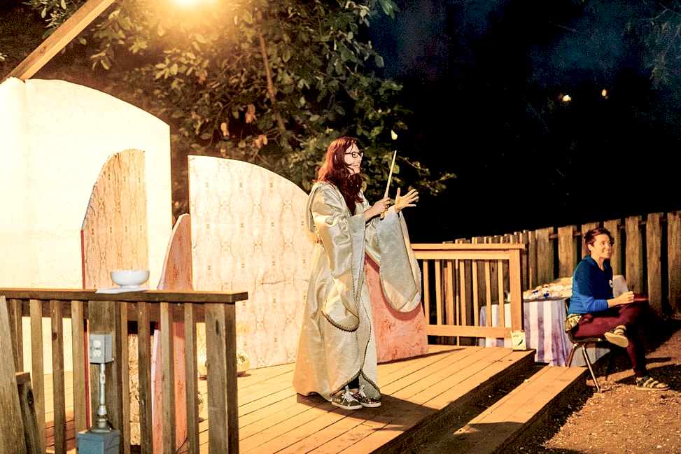 A woman in flowing robes, holding a wand, in front of the sunrise installation on a wood deck, with a sign language interpreter sitting nearby at the compact summer camp in la Honda, CA