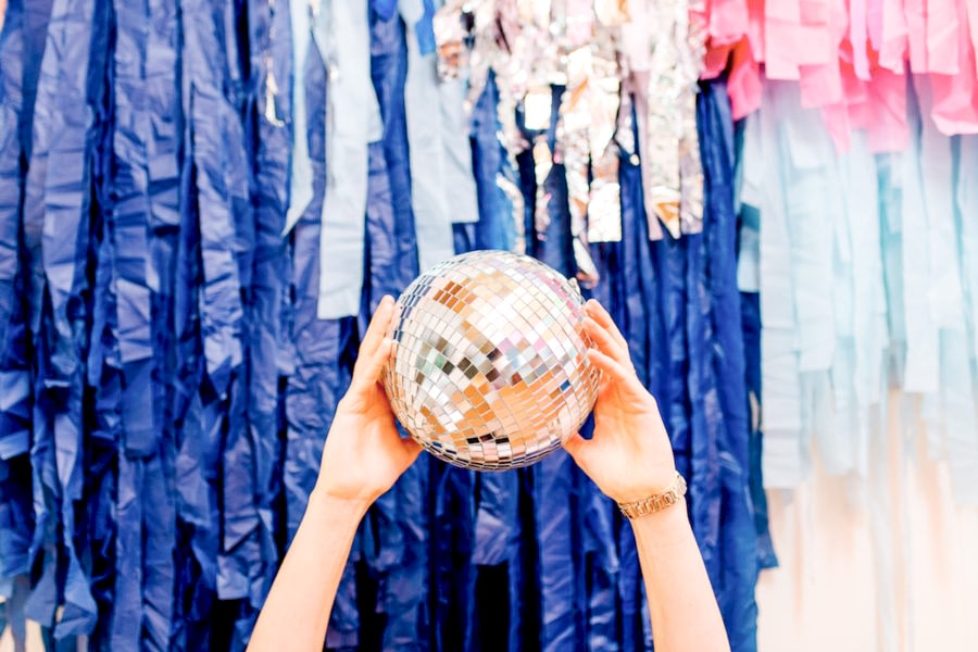 Two hands holding a medium-size disco ball in front of a colorful fringe installation