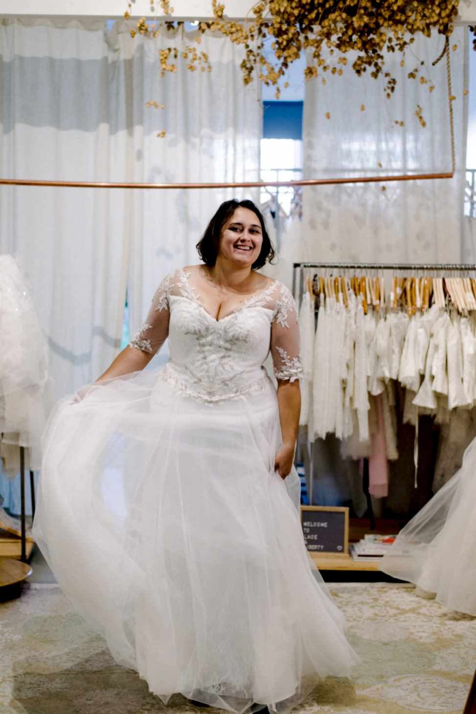 Plus size woman with short brunette hair smiles while twirling in wedding dress with lace sleeves, standing in front of a rack of plus size wedding dresses hanging on wooden hangers 