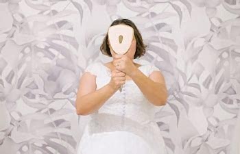 A plus size woman with chin-length dark hair wearing a wedding dress stands in front of a faded tropical leaf print papered wall, holding a hand mirror in front of her face