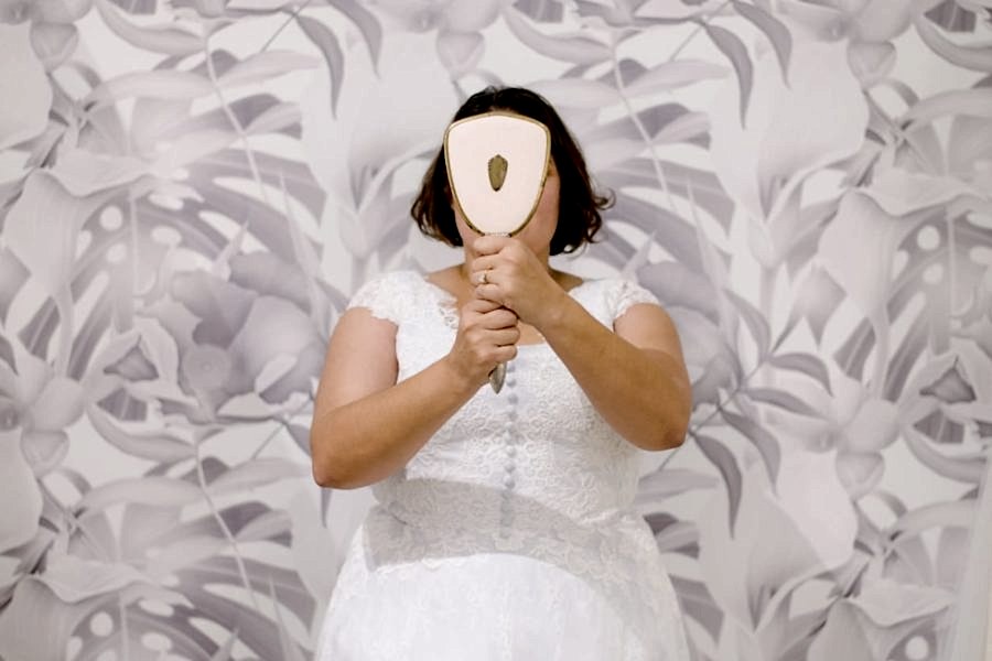 A plus size woman with chin-length dark hair wearing a wedding dress stands in front of a faded tropical leaf print papered wall, holding a hand mirror in front of her face