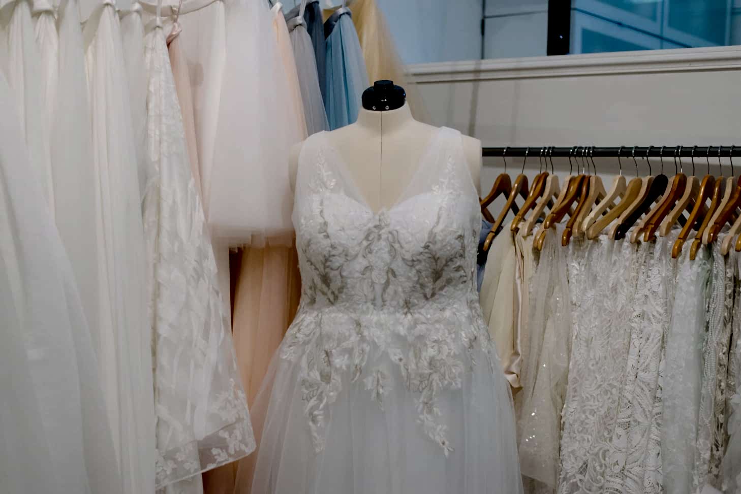 Plus size white lace wedding dress on mannequin in front of other plus size white lace wedding dresses hanging on brown wooden hangers
