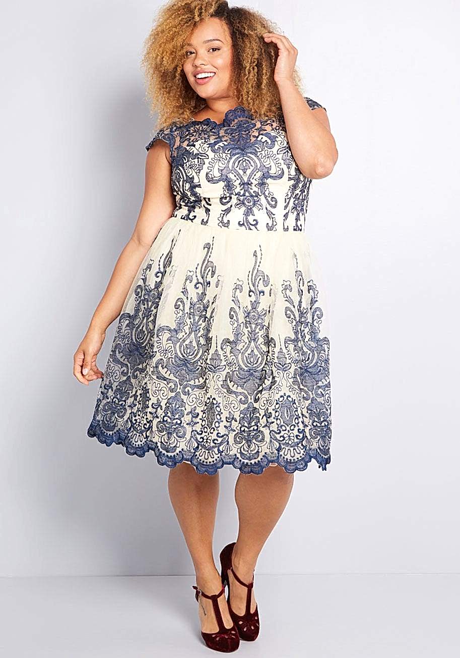 Black woman with blond curly hair wearing a white ankle length dress with blue lace details