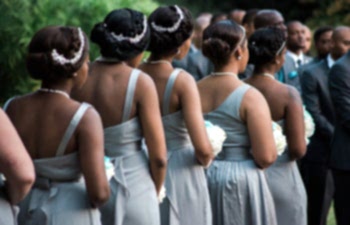 Five African-American bridesmaids with their hair up wearing light grey dresses and holding bouquets standing in a row with their backs to you during a wedding ceremony