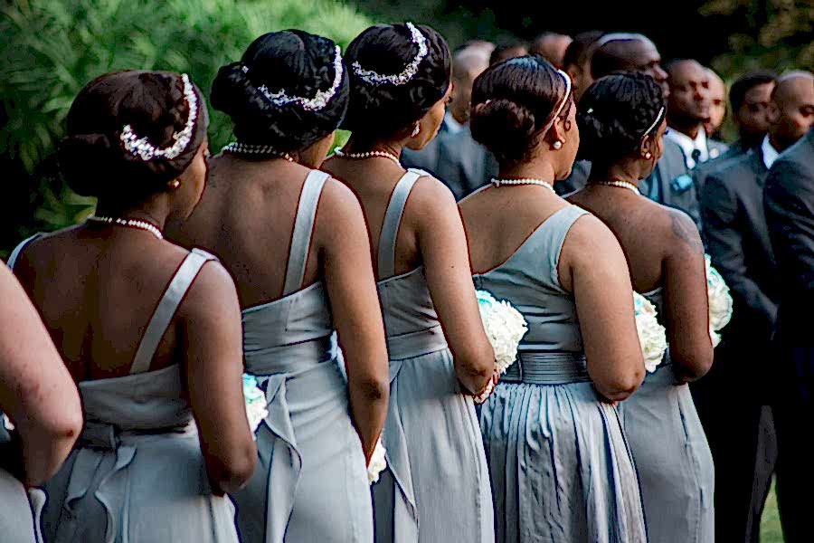 Five African-American bridesmaids with their hair up wearing light grey dresses and holding bouquets standing in a row with their backs to you during a wedding ceremony